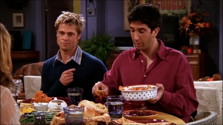 Brad Pitt as Will sitting next to Ross at the dinner table in a scene from Friends.