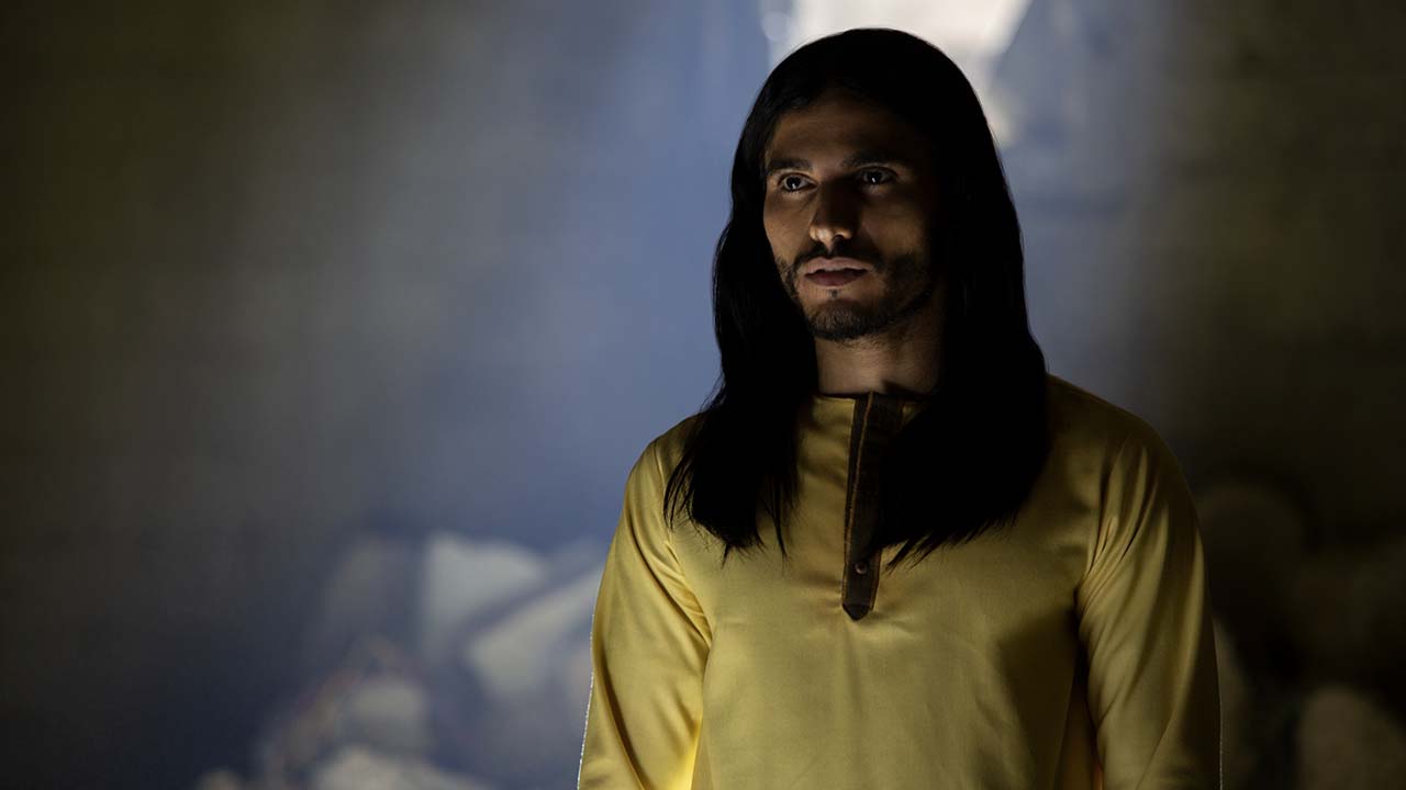 Um homem de cabelos compridos vestindo uma túnica amarela está de pé, a luz iluminando seu rosto em uma cena do Messias.