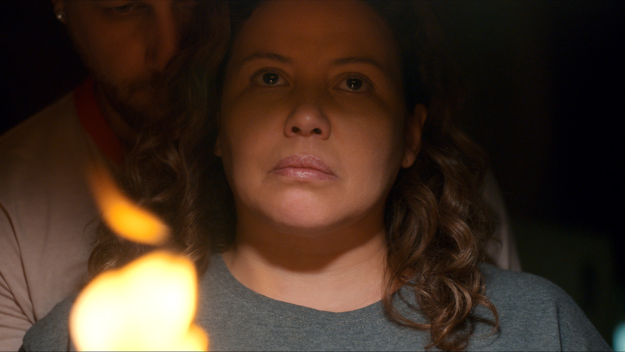 جاستینا ماچادو در نقش دولورس روچ در فیلم ترسناک دولورس روچ، در اتاقی تاریک به بالا نگاه می کند، شمعی پشت سرش.
