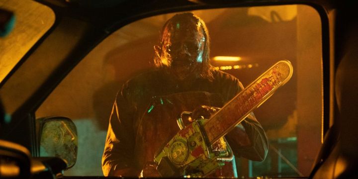 An old Leatherface holds his chainsaw standing next to a car
