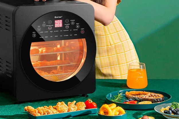The Costway 16-in-1 15-quart air fryer oven on a table with plates of food spread out around it.
