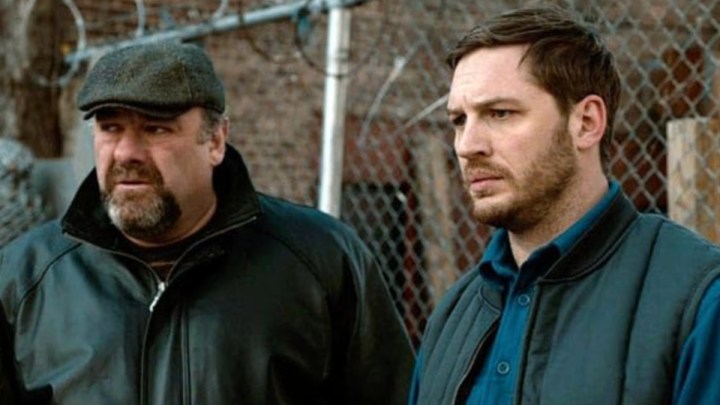 James Gandolfini and Tom Hardy stand next to each other in front of a fence in The Drop.
