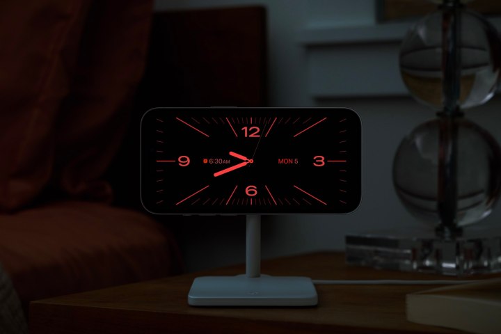 iPhone on charging stand showing red night mode analog clock in iOS 17 StandBy mode.