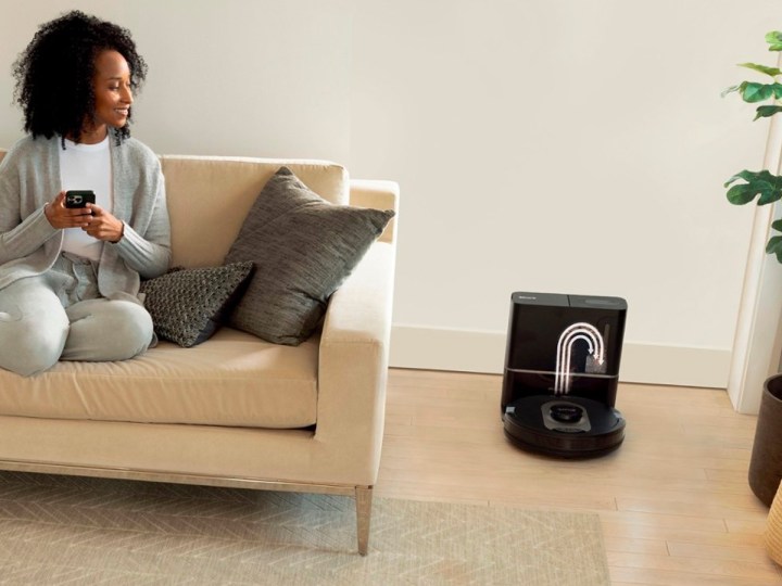 A woman on a sofa controls the Shark AI Ultra robot vacuum with her smartphone.