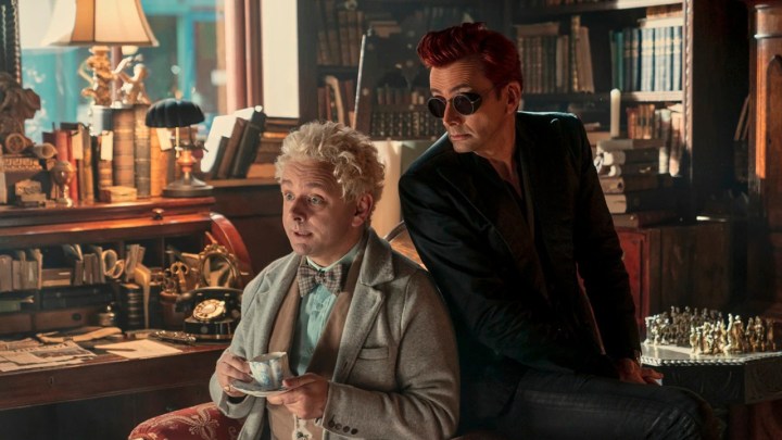 Michael Sheen sitting with a cup of tea and David Tennant sitting on the back of the chair behind him in a scene from Good Omens.