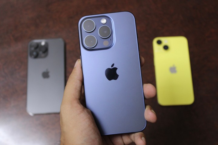 iPhone 15 Pro in mano con iPhone 14 Pro Max e iPhone 14 sullo sfondo.