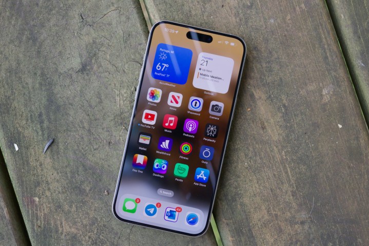 একটি iPhone 15 Pro Max, একটি কাঠের মেঝেতে শুয়ে আছে যার ডিসপ্লে চালু আছে।