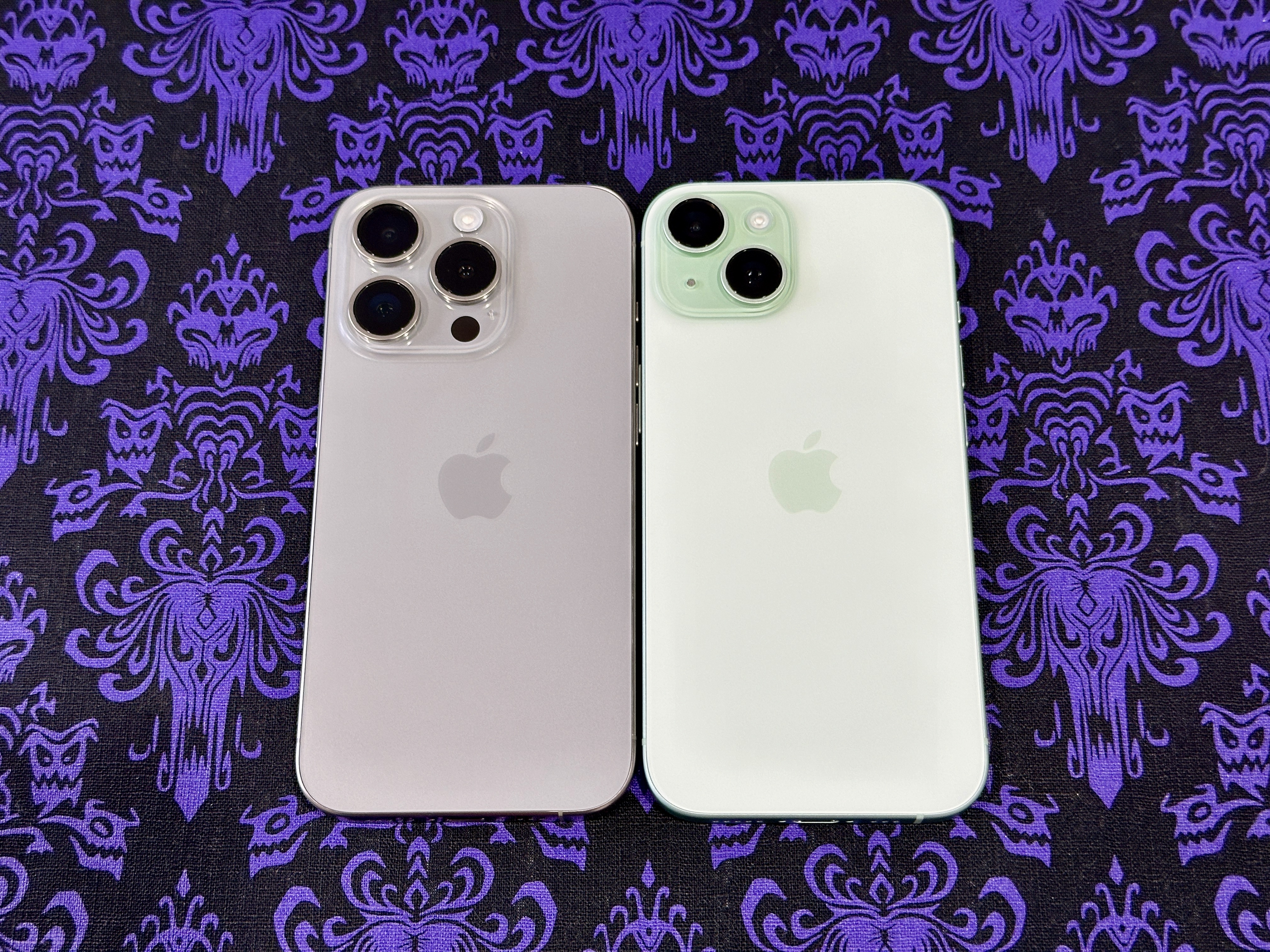 Натуральный титан iPhone 15 Pro (слева) и зеленый iPhone 15 на подставке для столовых приборов с обоями в Особняке с привидениями.