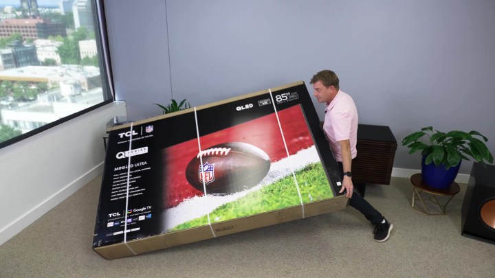 A man struggles to put an 85-inch boxed TV upright in a studio space. 