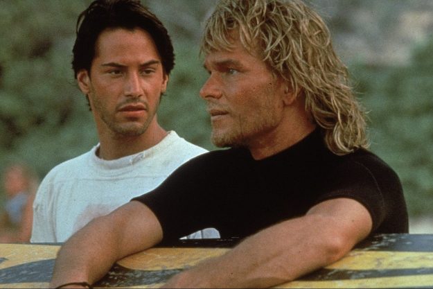 A man holds a surfboard while standing next to someone else in Point Break.