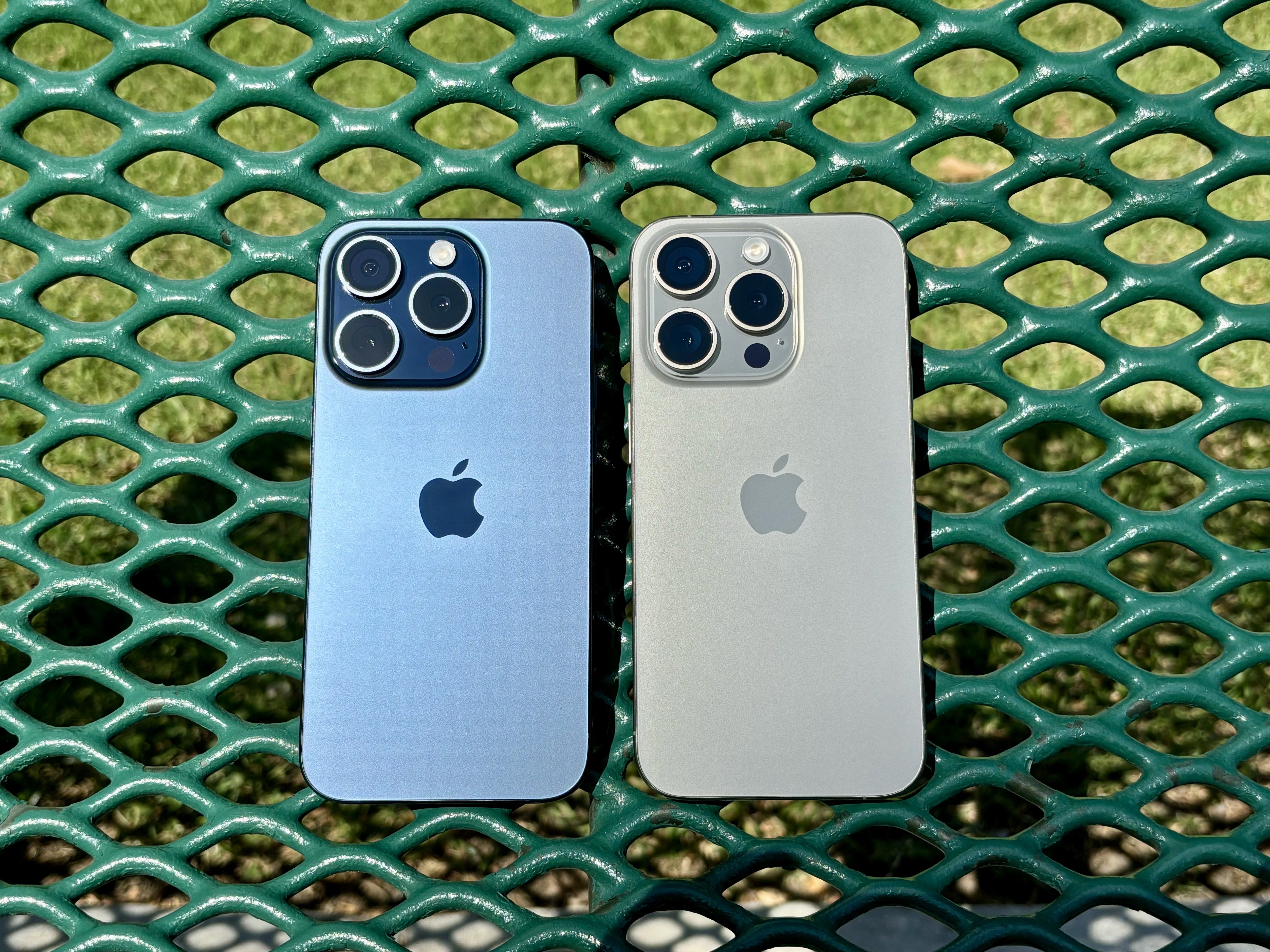 Mavi Titanyum (solda) ve Natural Titanium iPhone 15 Pro'lar yeşil bir park bankında.