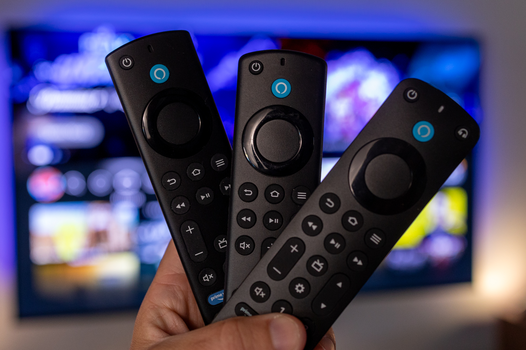 A trio of Amazon Fire TV remote controls held in a hand in front of a television.