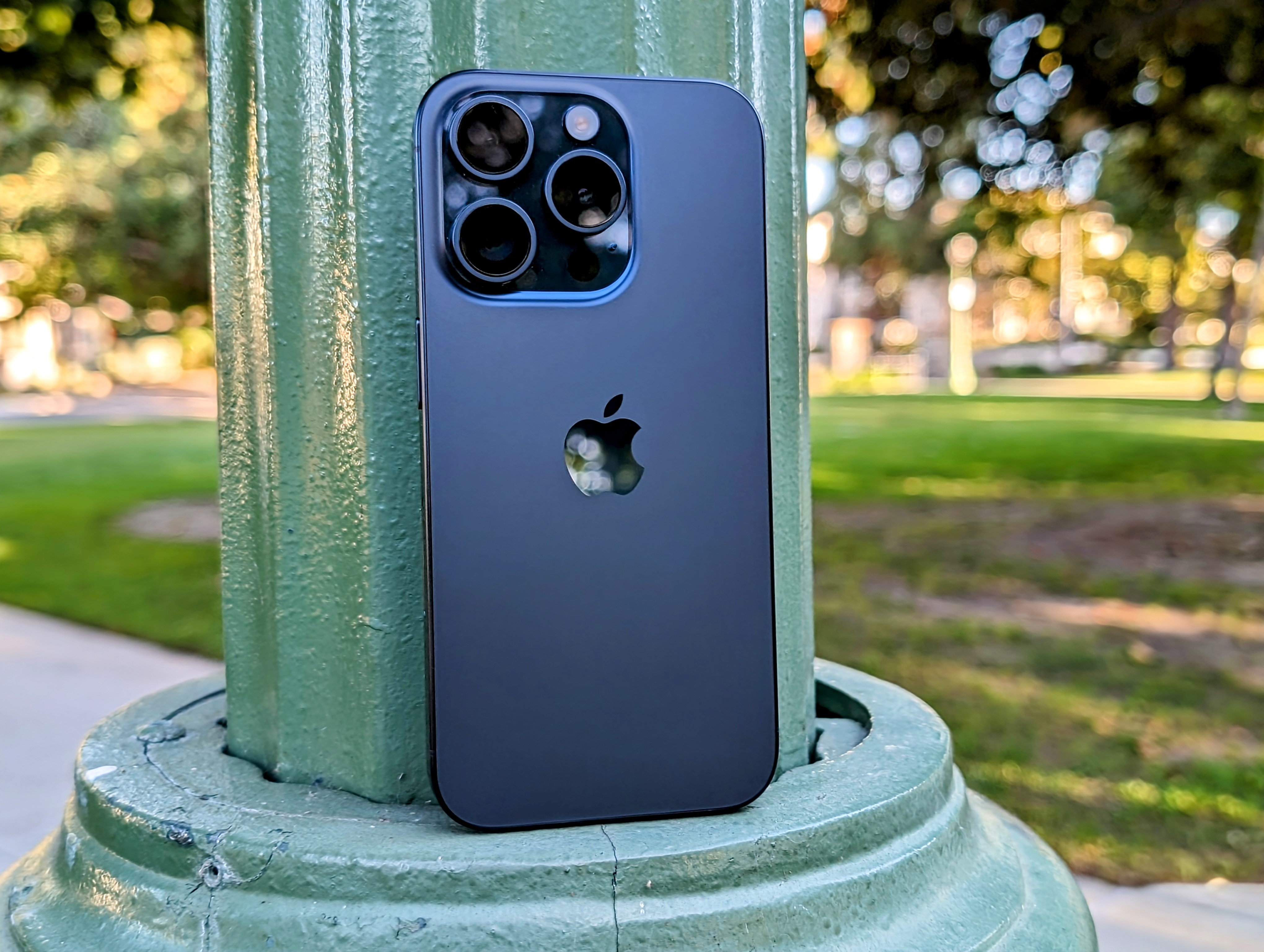 iPhone 15 Pro en titane bleu appuyé sur un lampadaire de parc.