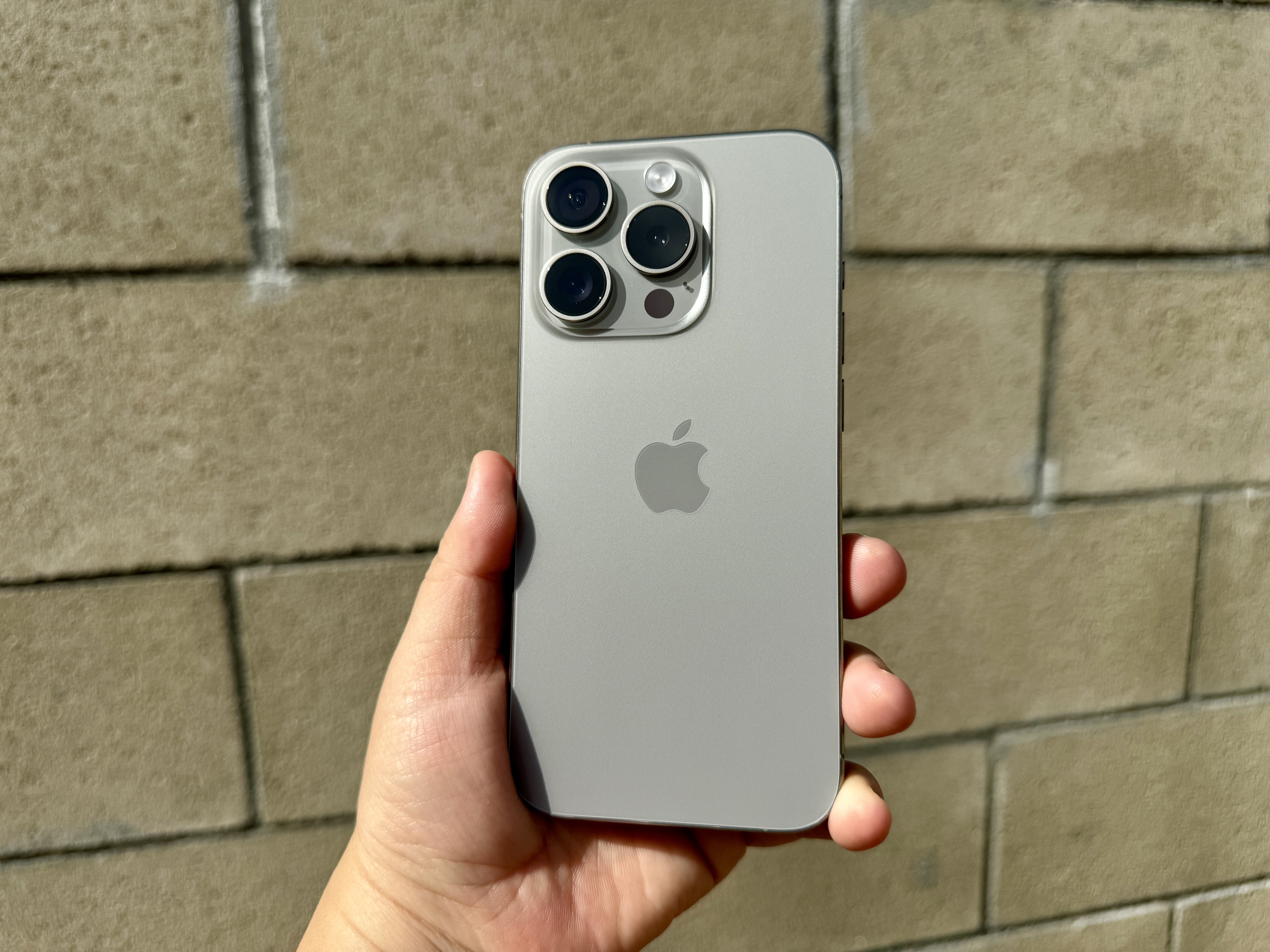 iPhone 15 Pro in Natural Titanium held in hand in front of a cement brick wall.