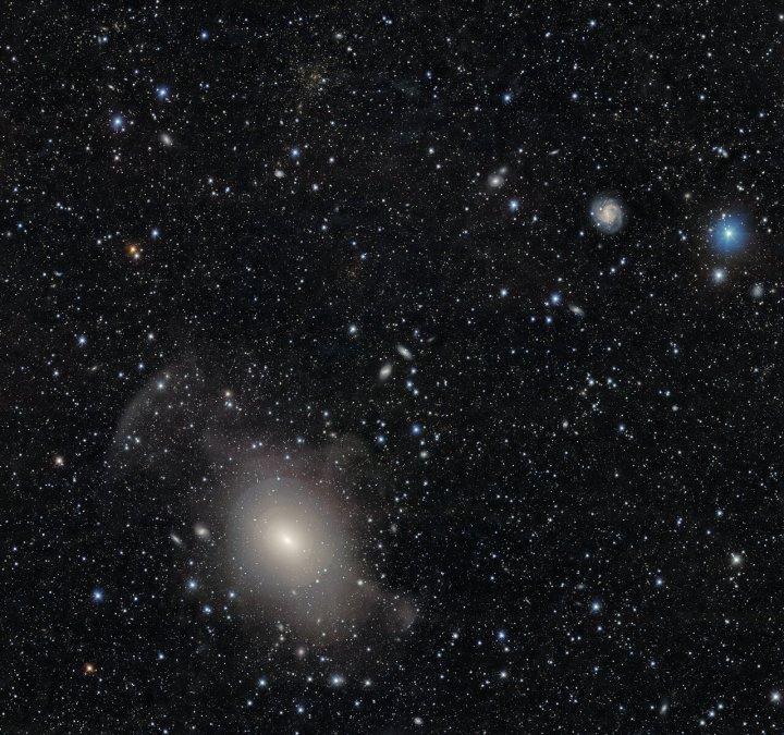 Las capas simétricas, parecidas a cebollas, de la galaxia de concha NGC 3923 se muestran en esta imagen rica en galaxias tomada por la Cámara de Energía Oscura del Departamento de Energía de los Estados Unidos (DOE) montada en el Telescopio de 4 metros Víctor M. Blanco de la Fundación Nacional de Ciencias (NSF) en el Observatorio Interamericano Cerro Tololo en Chile, un programa de NOIRLab de NSF. También se captura un cúmulo de galaxias masivo cercano que exhibe el fenómeno conocido como lente gravitacional.
