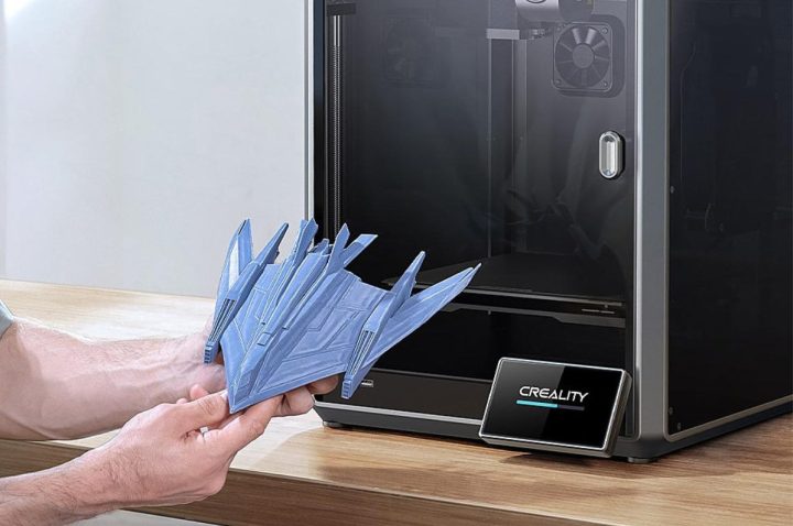A hand holds a 3D-printed spaceship in front of the Creality K1 3D printer.