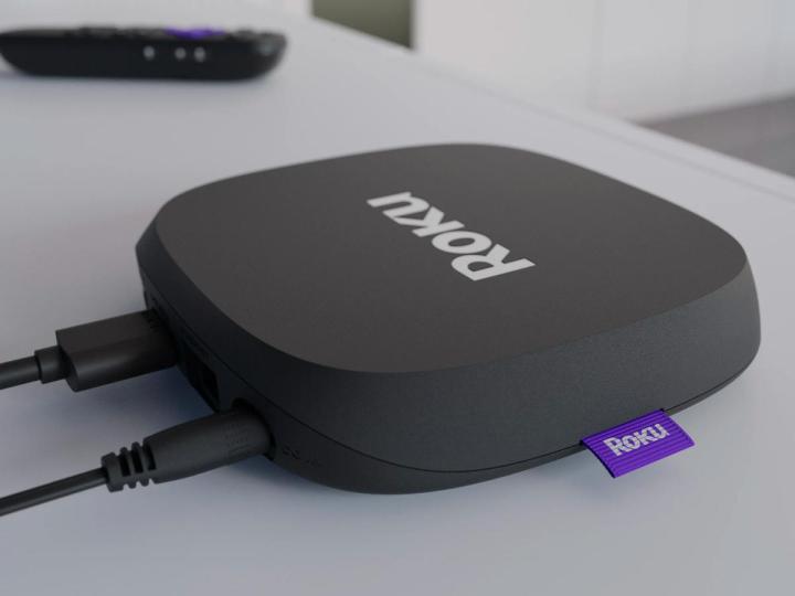 The Roku Ultra LT 4K streaming device on a media center with the included voice remote in the background.