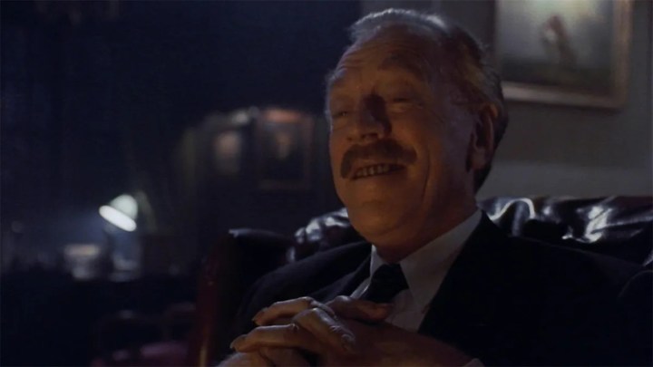 Un hombre mayor sentado en una silla sonriendo con aire de suficiencia en una escena de la película Needful Things.