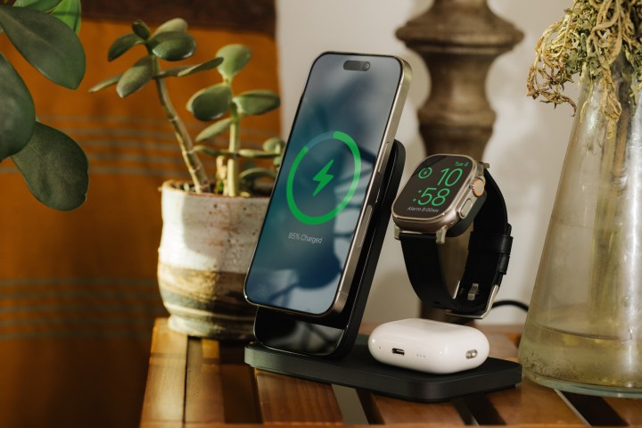 Black Nomad Stand One Max charging stand on household table with iPhone, Apple Watch Ultra, and AirPods Pro docked and charging.