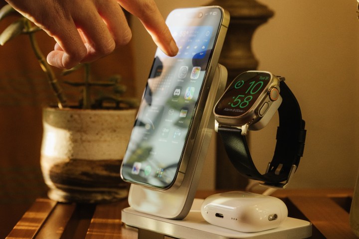 Person touching iPhone with one finger while docked on a Nomad Stand One Max charging stand sitting on a household table.