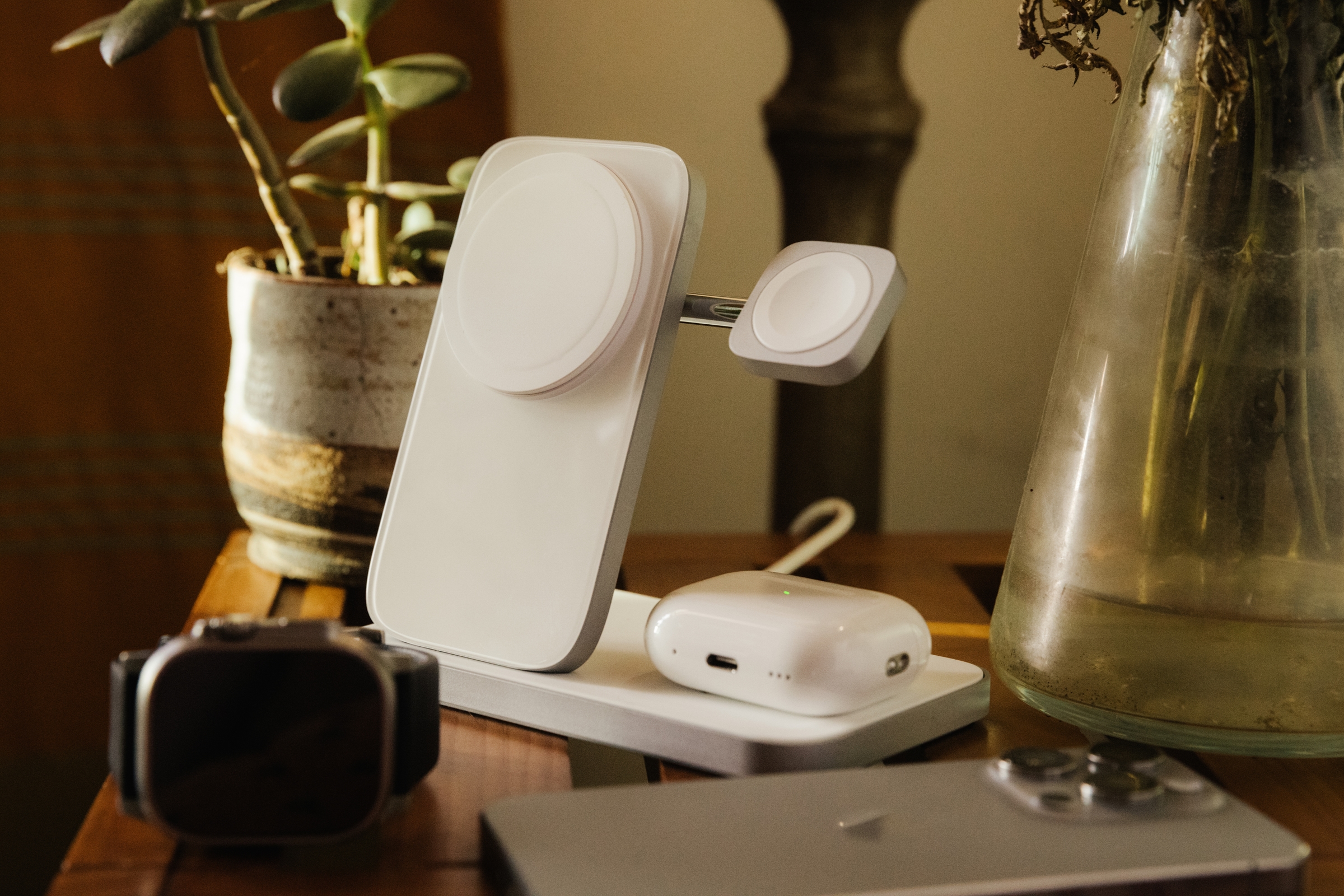 Silver Nomad Stand One Max iPhone charging stand on table with AirPods Pro, iPhone, and Apple Watch lying in front.