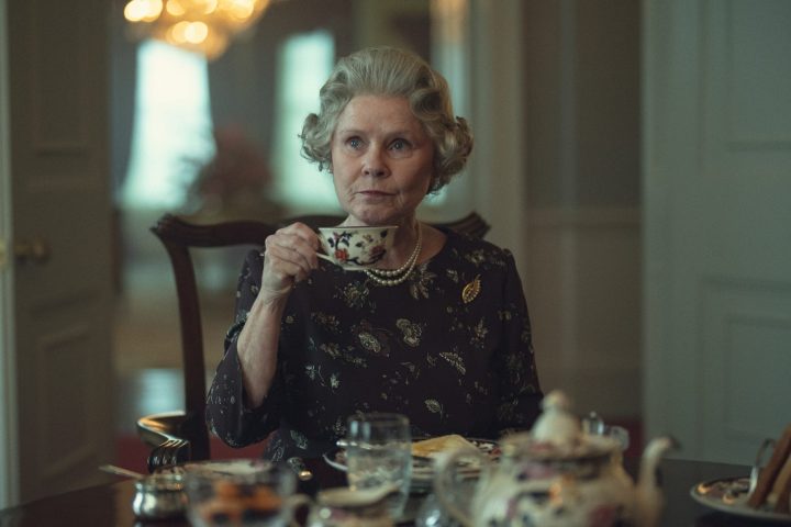 Queen Elizabeth sits a table and holds up her tea cup.