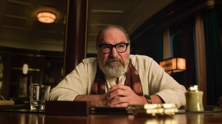 Mandy Patinkin sentada en un escritorio, con las manos entrelazadas frente a él en una escena de Muerte y otros detalles.