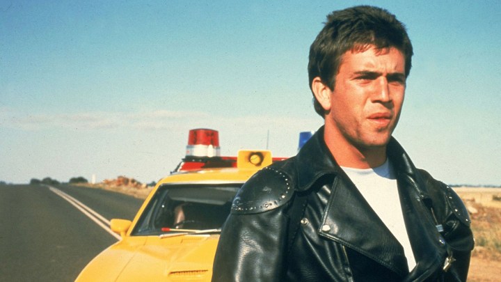 Mel Gibson beside a car standing in an empty road in a scene from Mad Max.