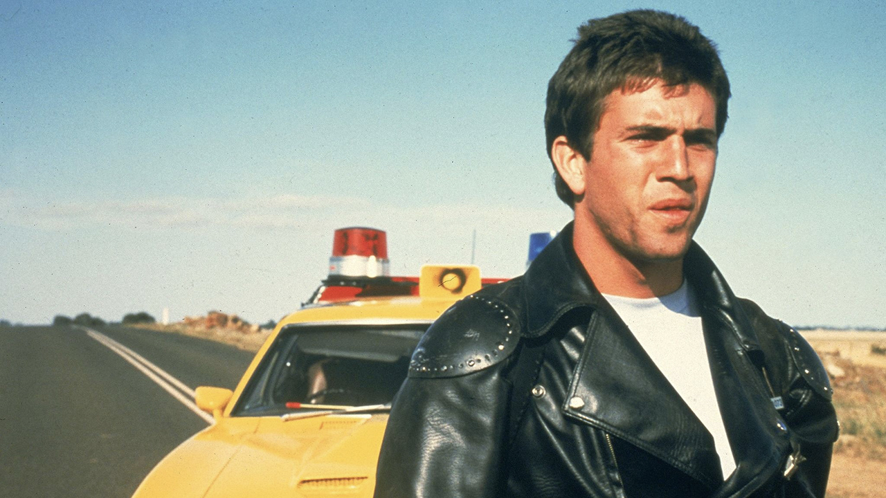 Mel Gibson ao lado de um carro parado em uma estrada vazia em uma cena de Mad Max.
