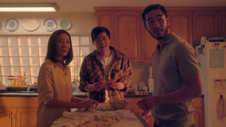 Two sons and their mom in the kitchen all looking at something in a scene from The Brothers Sun on Netflix.