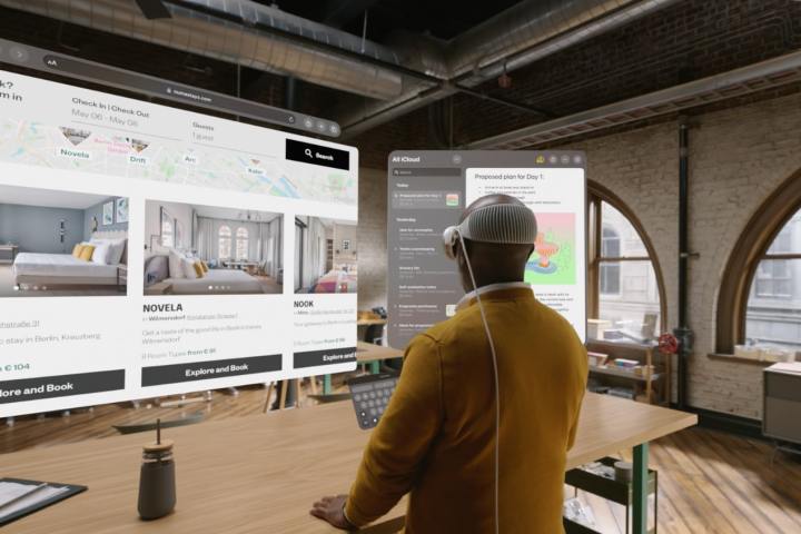 A person uses Apple's Vision Pro headset at a desk. They are typing on a virtual keyboard, while two virtual monitors float in the air in front of them.