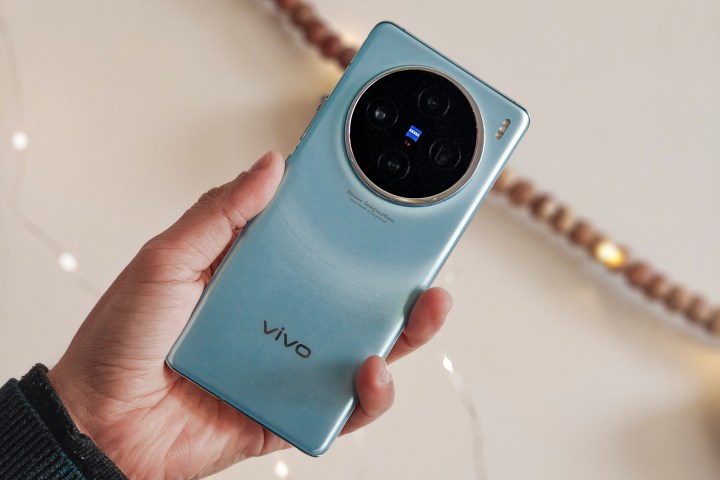 Vivo X100 Glacier Blue held in hand against fairy lights and garland of wooden beads.