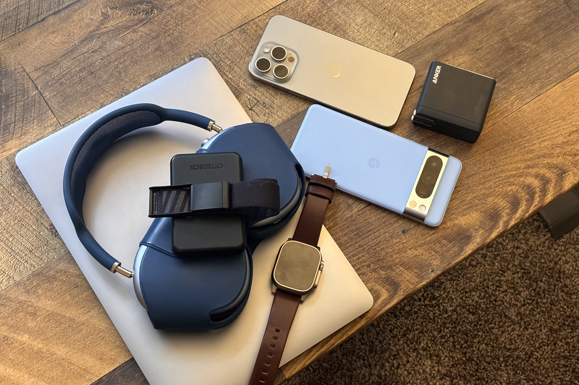 A MacBook, AirPods Max, Apple Watch, iPhone, Google Pixel, and Anker charger all lying next to each other on a desk.