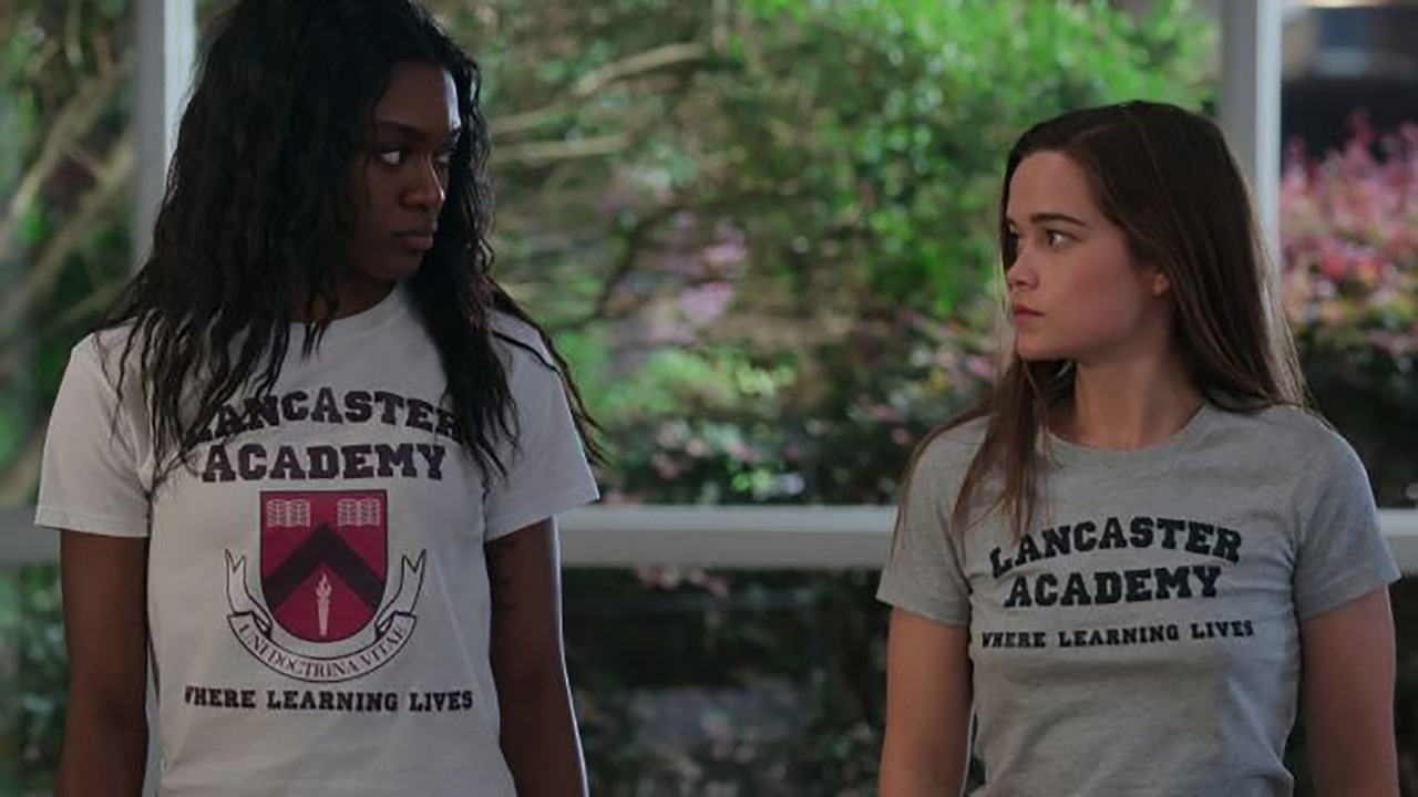 नेटफ्लिक्स पर फर्स्ट किल के एक दृश्य में दो युवा महिलाएं एक-दूसरे को देख रही हैं, दोनों ने हाई स्कूल टी-शर्ट पहनी हुई है।