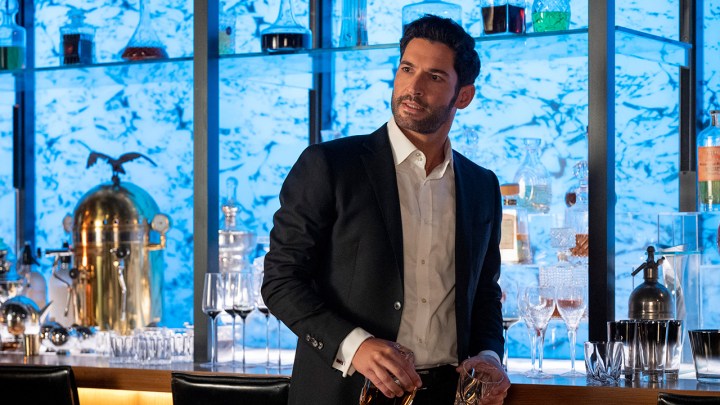 Tom Ellis as Lucifer Morningstar standing by his bar in a suit, looking at someone in a scene from Lucifer on Netflix.