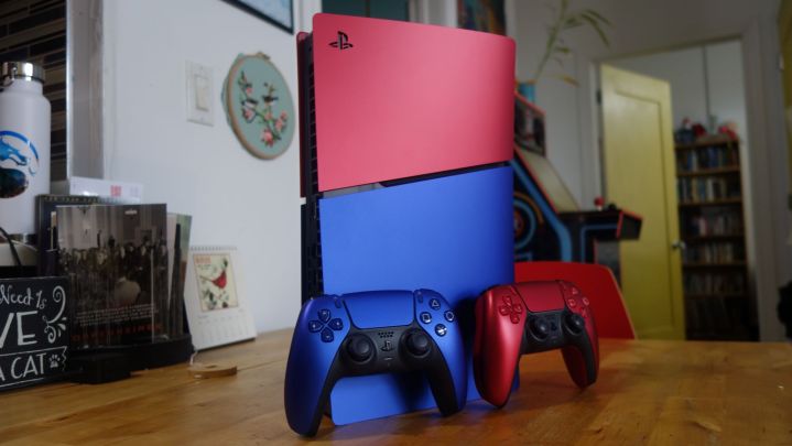 A red and blue PS5 stands on a table with matching controllers.