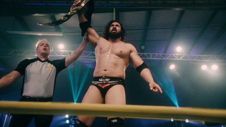 A man holding up the arm of a wrestler in the ring who is holding a belt in a scene from the Netflix series Wrestlers.