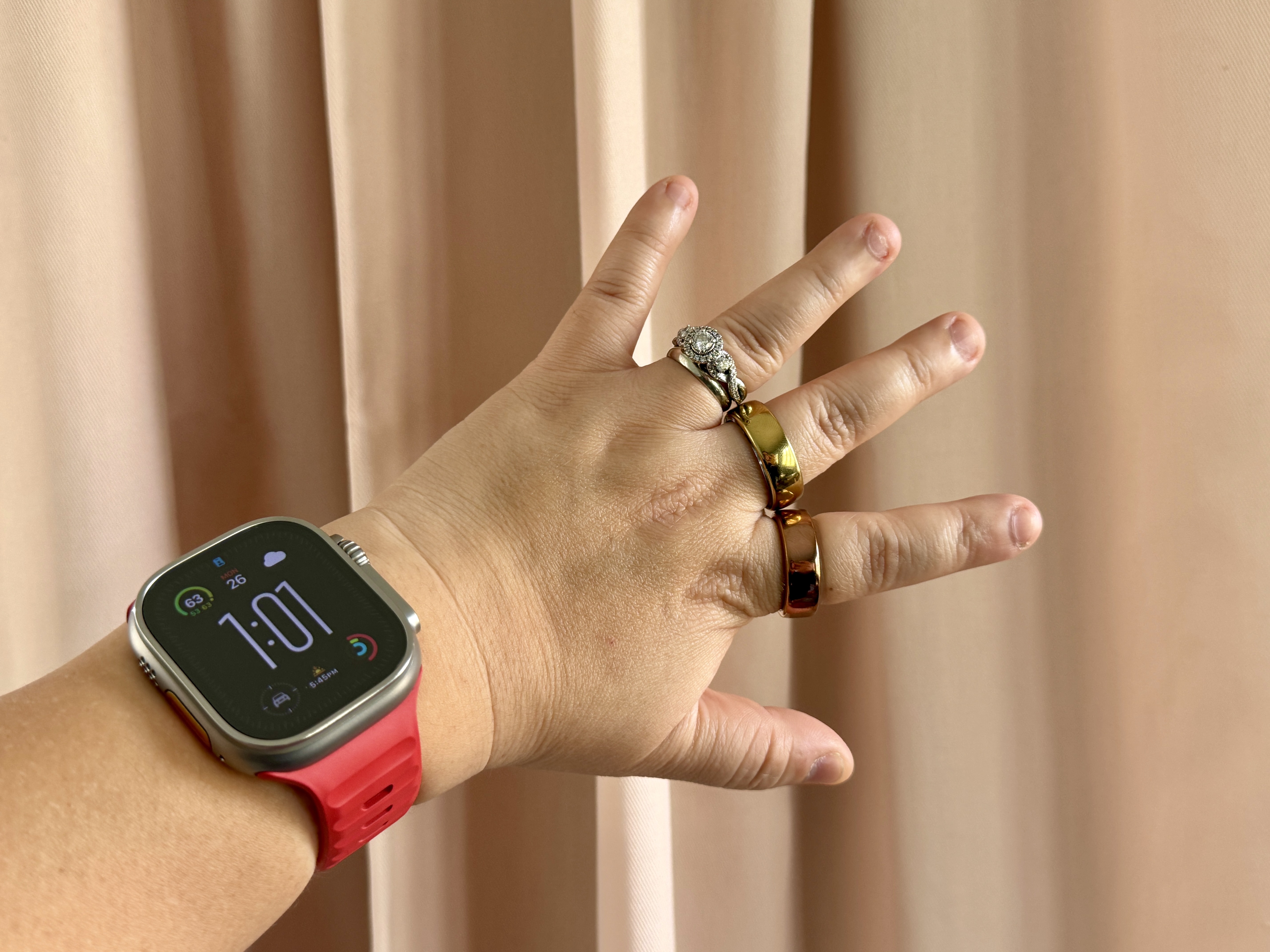 An Apple Watch Ultra with red strap on wrist with a Movano Evie and Oura Ring on fingers.