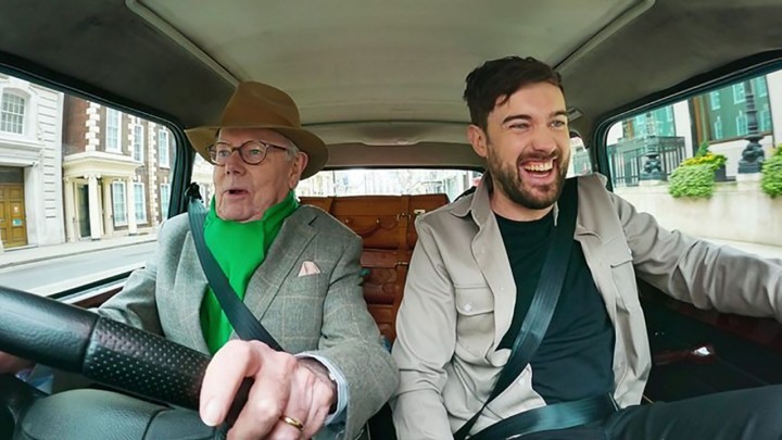 Jake Whitehall in a car with his father Michael driving, his father looking terrified and Jack smiling in a scene from Jack Whitehall: Travels with My Father on Netflix.