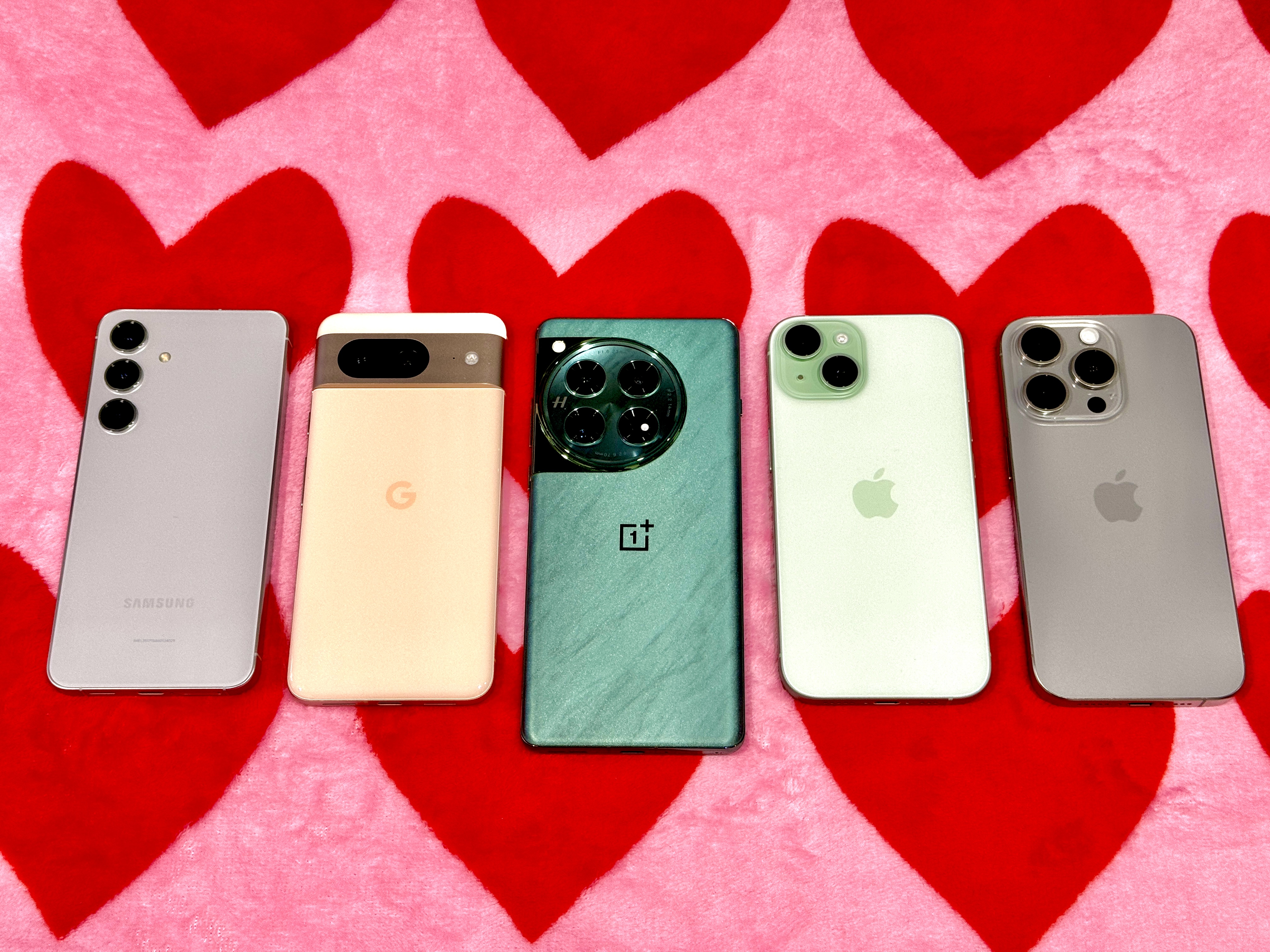 Samsung Galaxy S24 خاکستری (سمت چپ)، رزگلد Google Pixel 8، Flowy Emerald OnePlus 12، Green iPhone 15، Titanium Grey iPhone 15 Pro روی یک پتو قلبی صورتی و قرمز.