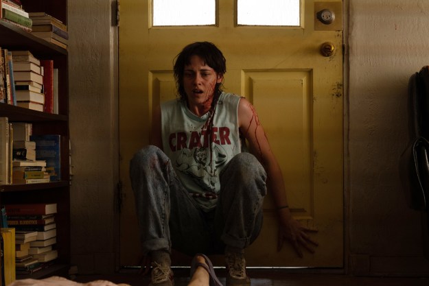 A blood-splattered Kristen Stewart sits against a door in Love Lies Bleeding.