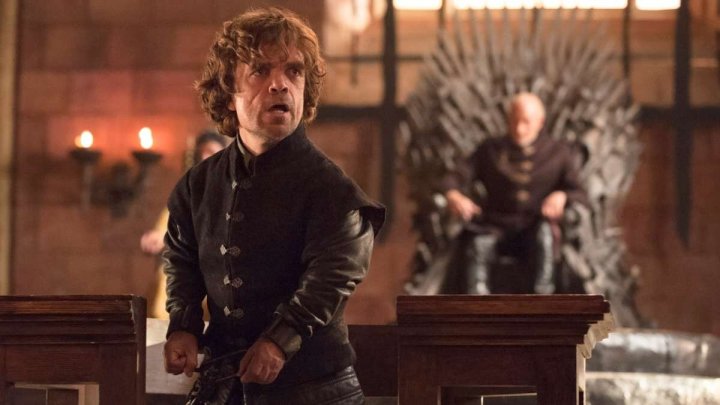 Peter Dinklage as Tyrion Lannister talking while his father sits on the Iron Throne behind him in Game of Thrones.