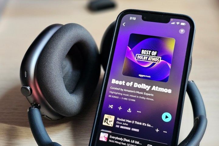 一部 iPhone 14，显示 Amazon Music 应用程序和杜比全景声 (Dolby Atmos) 音乐播放列表，旁边是一组 Apple AirPods Max 耳机。