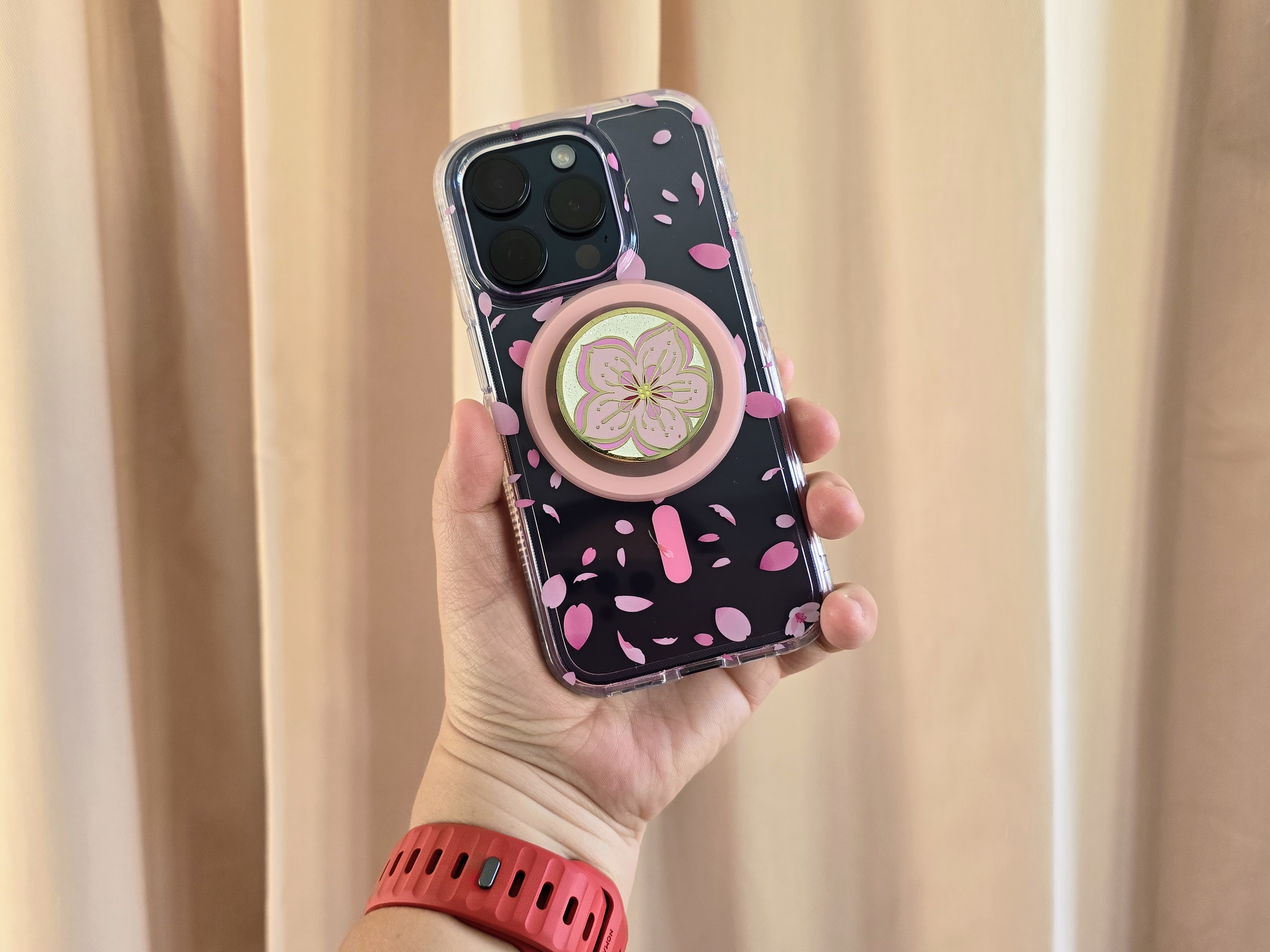 A Titanium Blue iPhone 15 Pro in a Tech21 Cherry Blossom case with a cherry blossom PopSocket MagSafe grip attached.