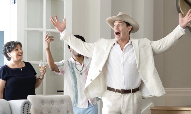 John Cena in a white suit and cowboy hat, his arms spread open and mouth wide in a scene from Ricky Stanicky.