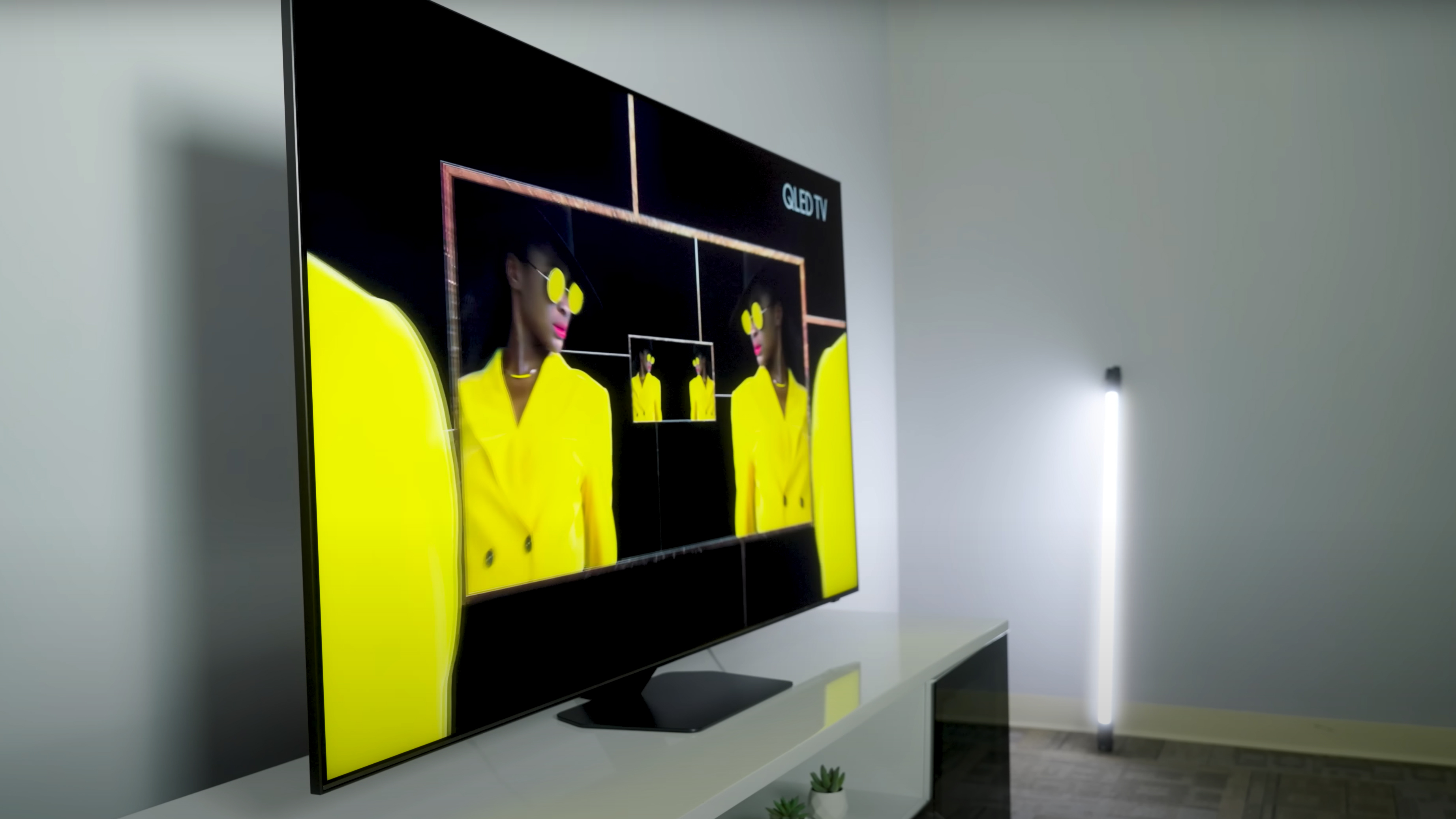 An editorial image of a woman in neon yellow suit and glasses reflected infinitely in a mirror on a Samsung QN90D TV.