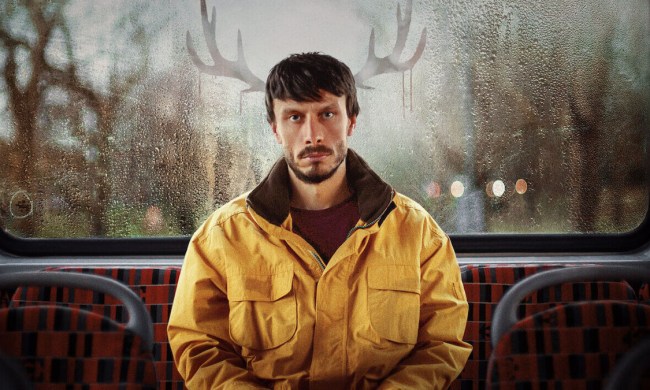 Richard Gadd in Baby Reindeer sitting on a bus with a yellow coat staring forward, the image of deer ears on either side of him.