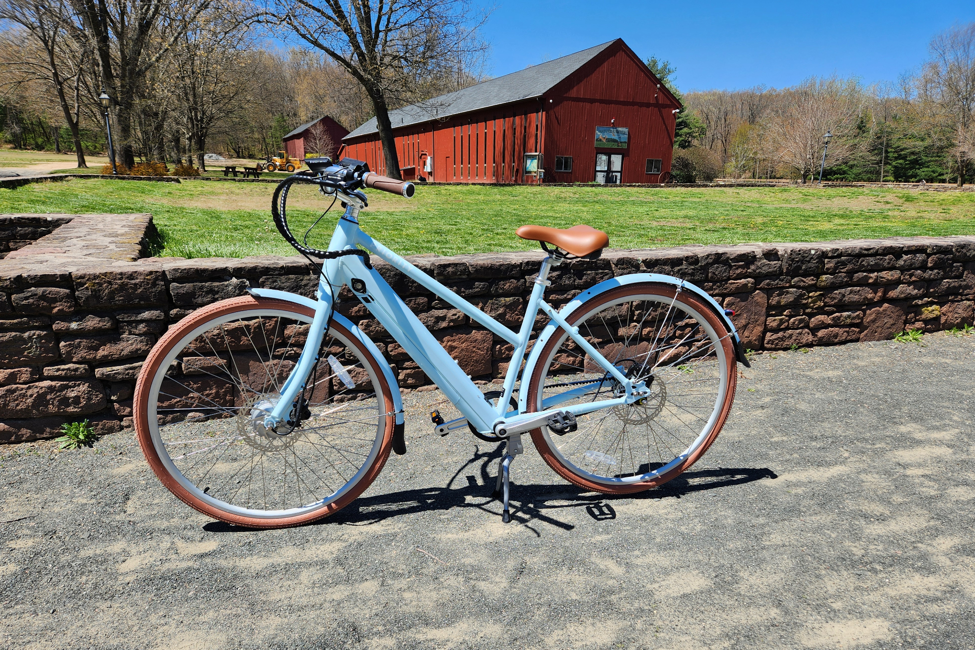 Priority Bicycles e-Classic Plus 电动自行车左侧面照片停在石墙前的碎石上，背景是一个红色改建的谷仓。