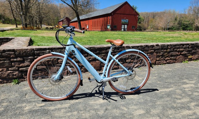 Priority Bicycles e-Classic Plus e-bike left profile shot parked on crushed stone in front of a stone wall with a red converted barn in the background.