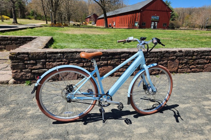 Priority Bicycles e-Classic Plus 电动自行车右侧轮廓停在石墙前的碎石上，背景是红色改建的谷仓。
